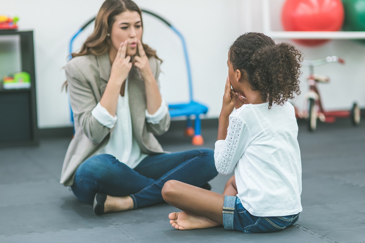 A female speech pathologist is teaching a child patient how to speak by having her touch her mouth