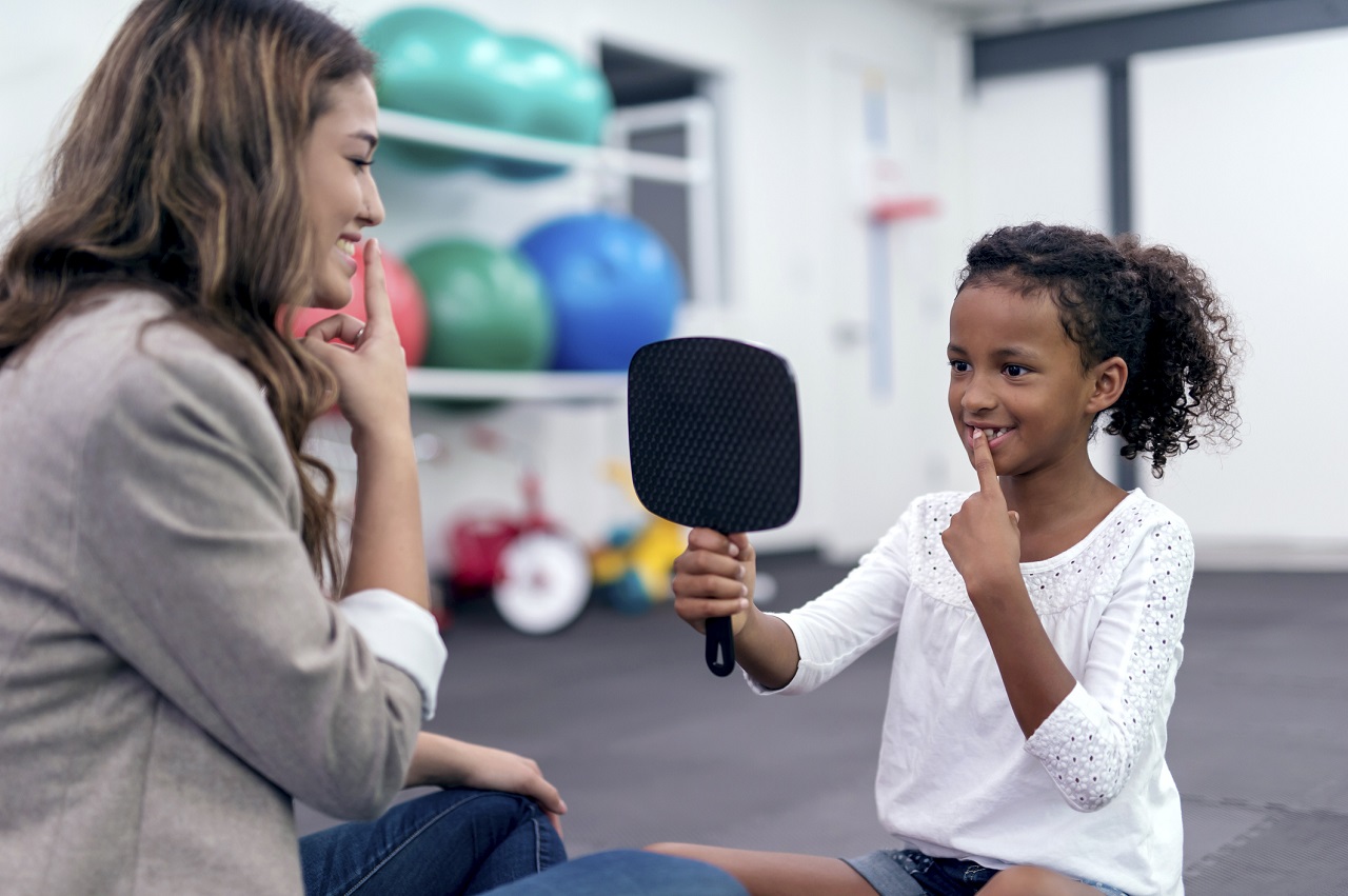 Cute African American girl using a mirror for speech therapy with the guidance of a speech therapist